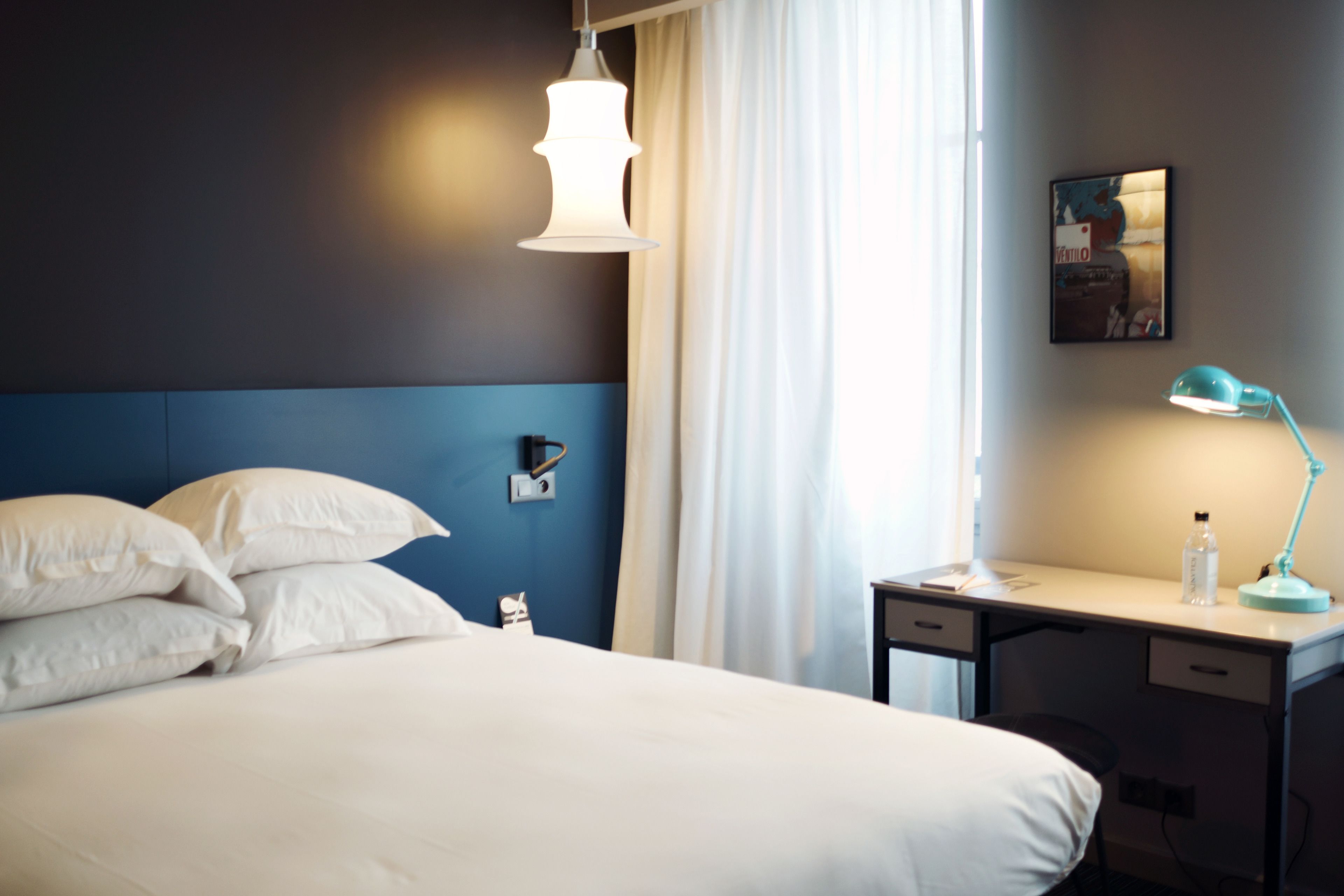 A hotel room with a bed, a desk, a lamp, a picture on the wall, and a window with curtains.