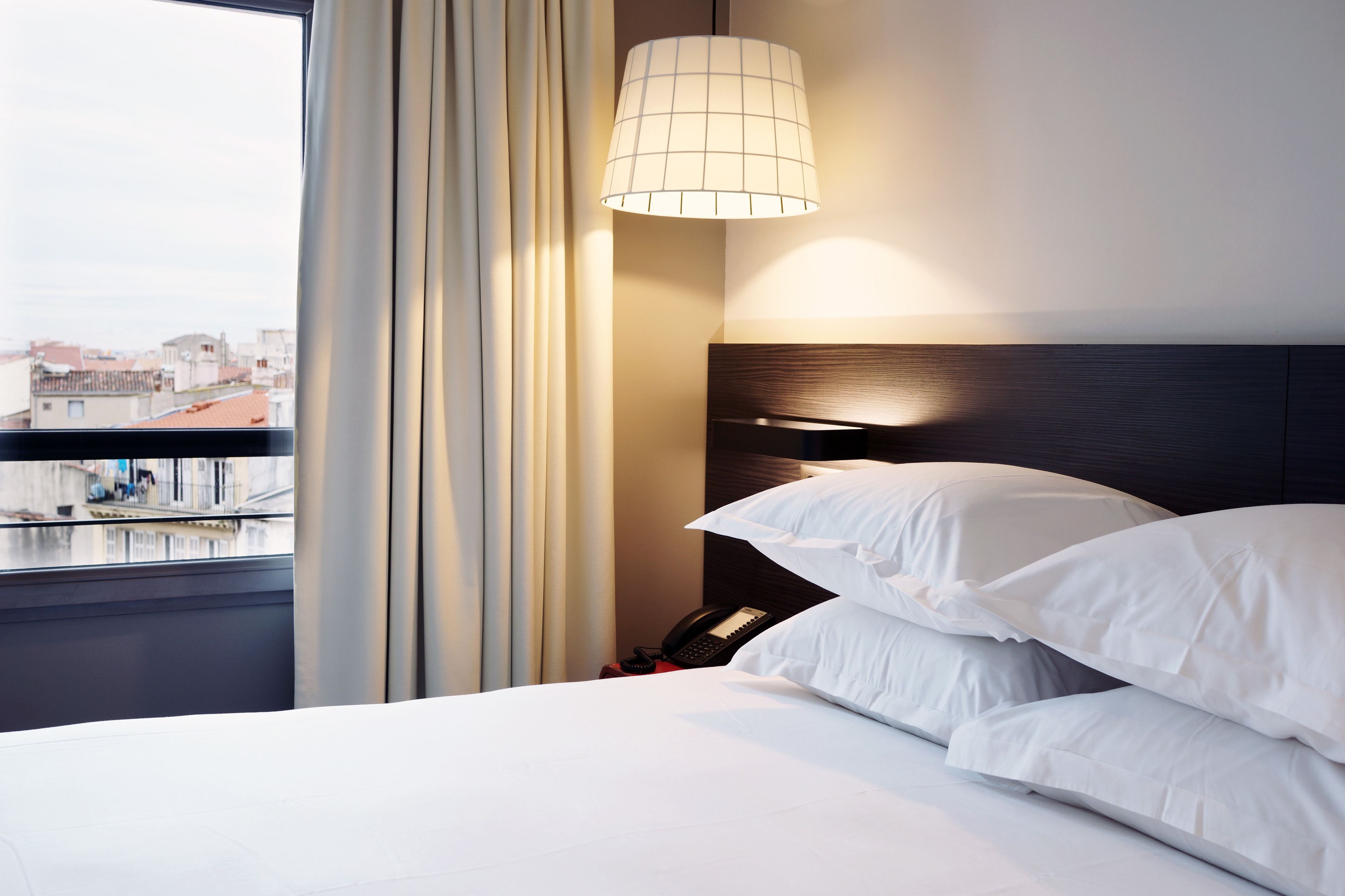 Ein Hotelzimmer mit einem Bett, einem Nachttisch, einer Lampe und Blick auf die Stadt durch ein Fenster.