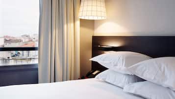 Ein Hotelzimmer mit einem Bett, einem Nachttisch, einer Lampe und Blick auf die Stadt durch ein Fenster.