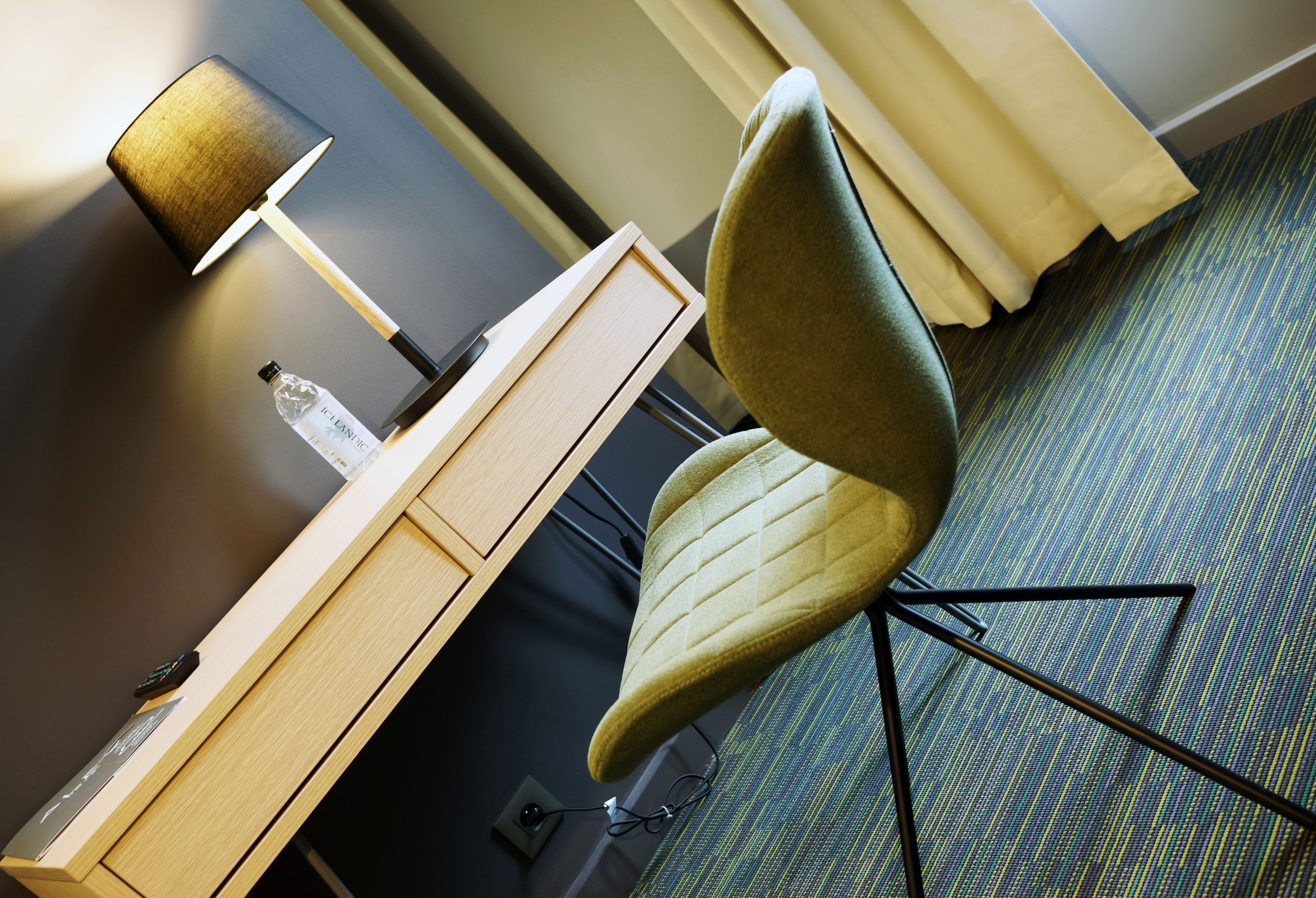A hotel room with a wooden desk, a lamp, a bottle, and a green patterned chair.