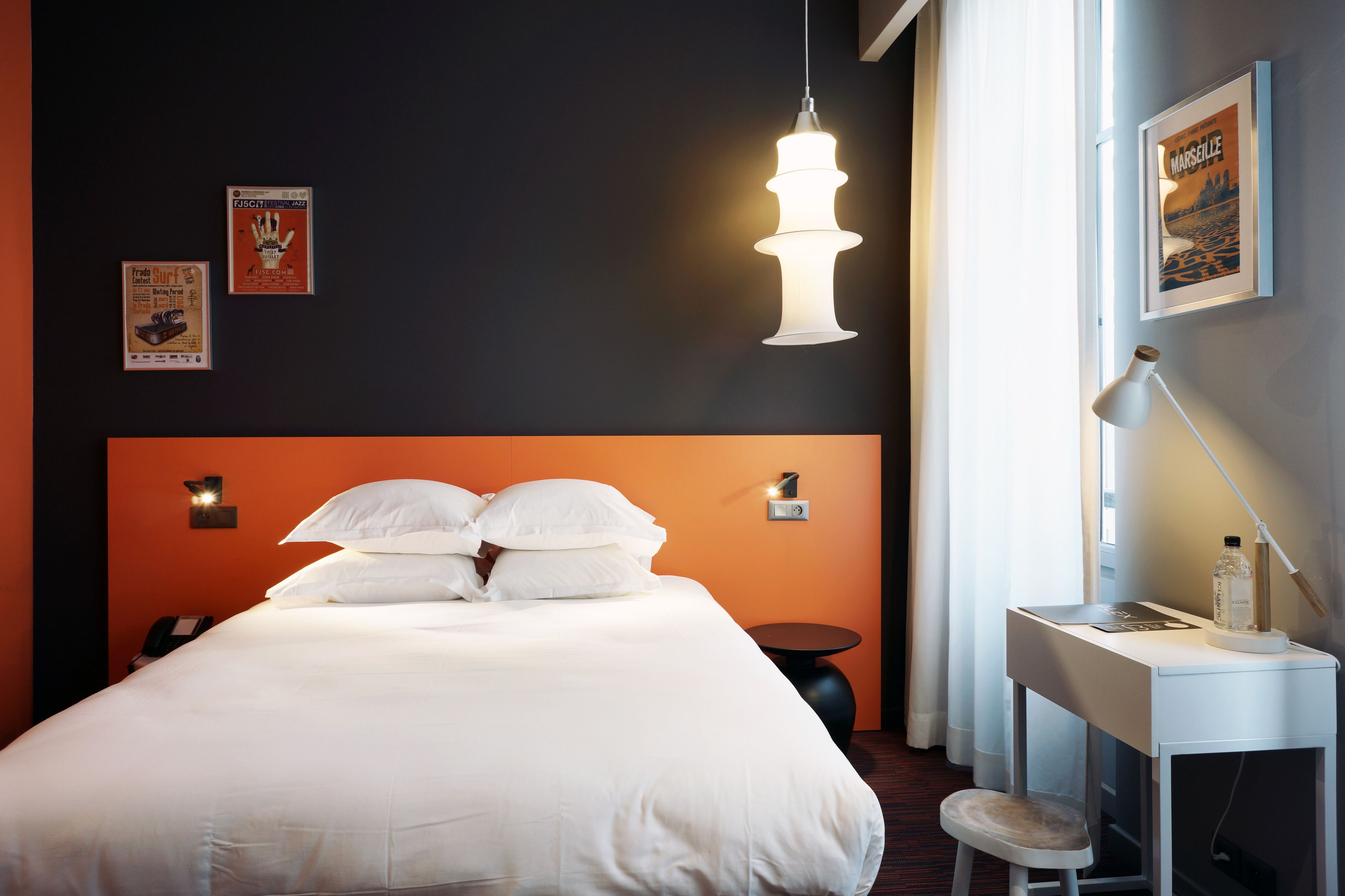 A hotel room with a large bed, a desk with a chair, a hanging light fixture, and a framed picture on the wall.