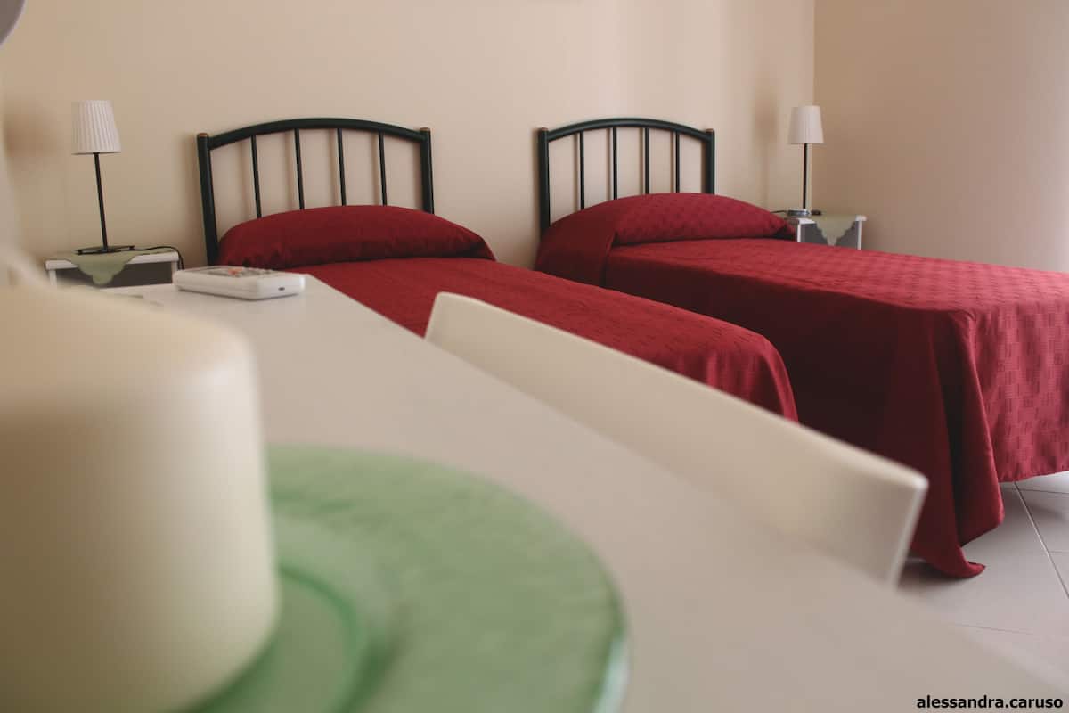 A hotel room with two beds, each with a red bedspread and black headboards. There are bedside tables with lamps and a small table with a remote control.