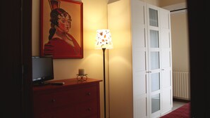 Une pièce avec une télévision posée sur une commode rouge, une lampe avec un abat-jour à motifs et une armoire blanche avec des portes en verre.