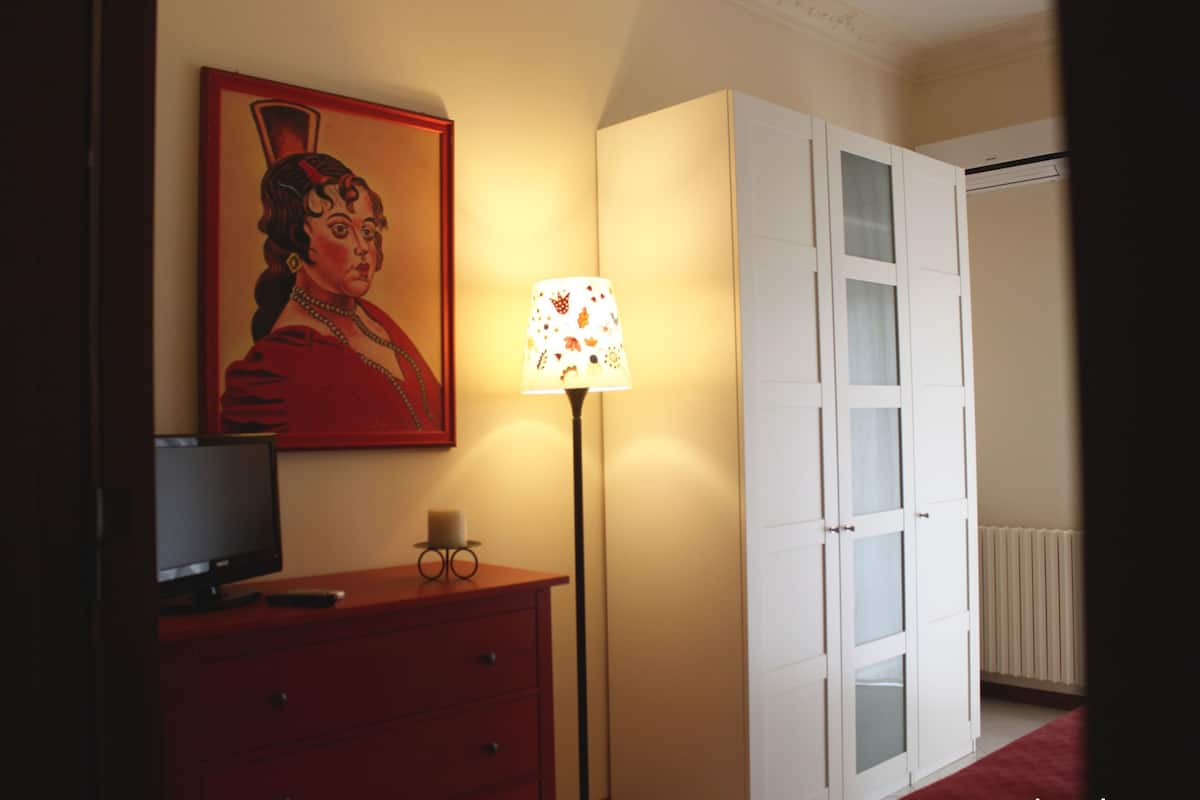 A room with a television on a red dresser, a lamp with a patterned shade, and a white wardrobe with glass doors.