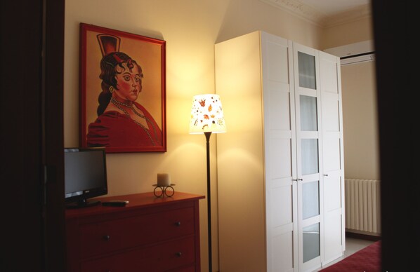 Una habitación con una televisión sobre un aparador rojo, una lámpara con pantalla estampada y un armario blanco con puertas de cristal.