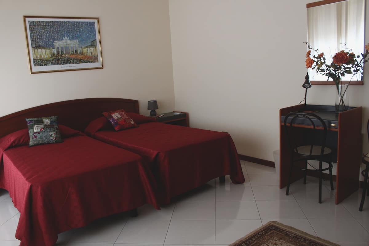 A room with two beds, a desk, a chair, a lamp, a vase with flowers, a framed picture on the wall, and a rug on the floor.