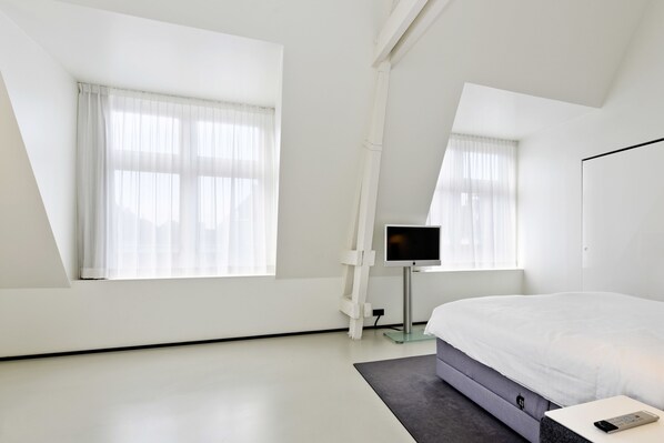 A modern bedroom with a large bed, a TV on a stand, and a window with sheer curtains. - Urban Residences Maastricht (Maastricht)