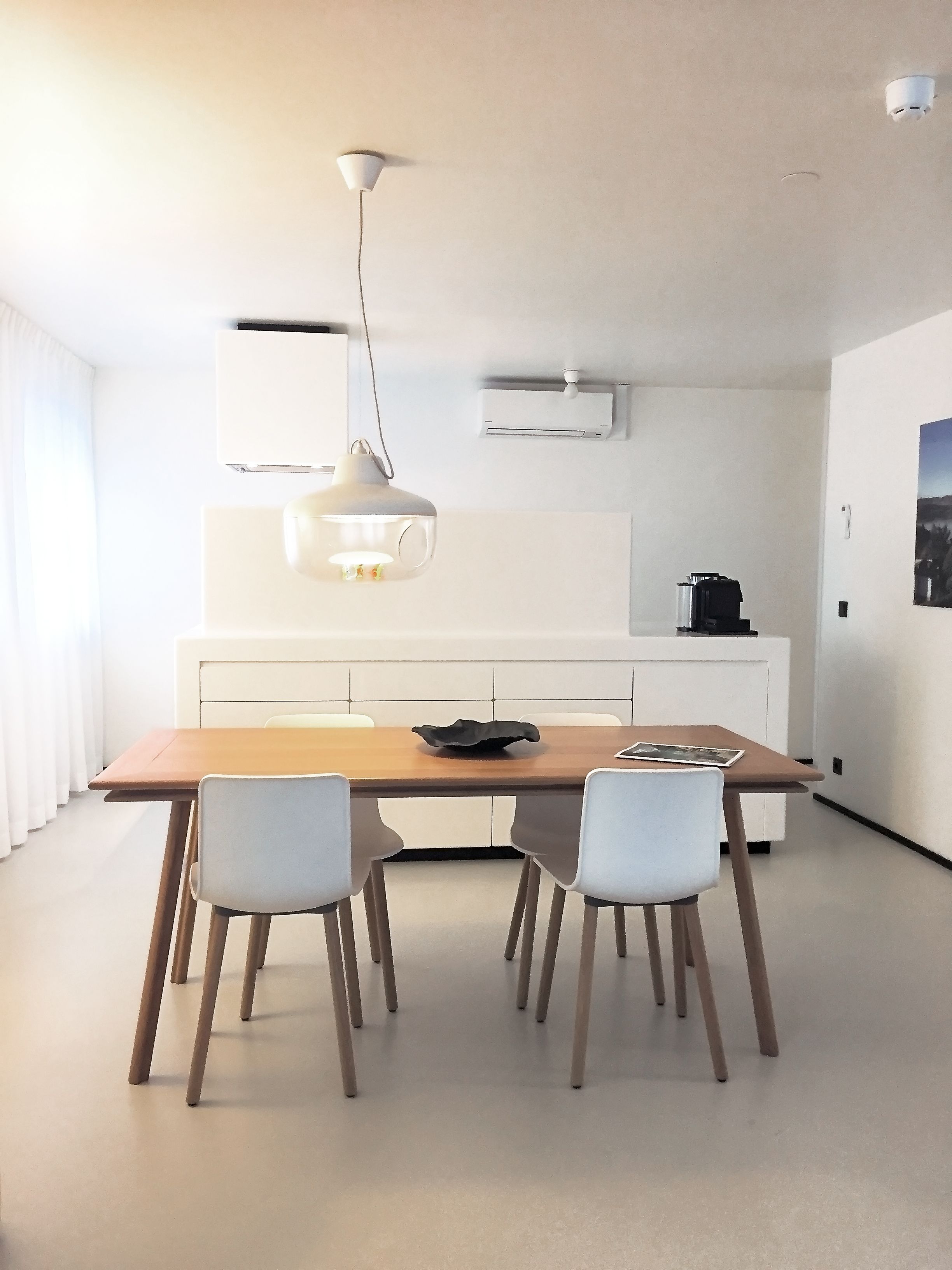 A modern kitchen with a wooden table, two white chairs, a pendant light, and white cabinetry.