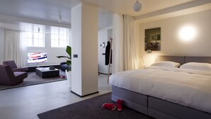 Una habitación de hotel moderna con una cama grande, una zona de estar con un sofá y una mesa de centro, un televisor y una planta en maceta.