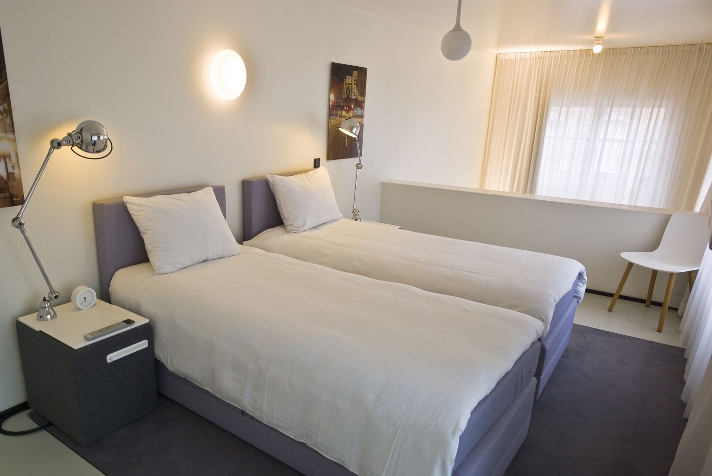 Ein Hotelzimmer mit zwei Betten, einem Nachttisch mit Lampe, einem Sessel und einem Fenster mit Vorhängen.