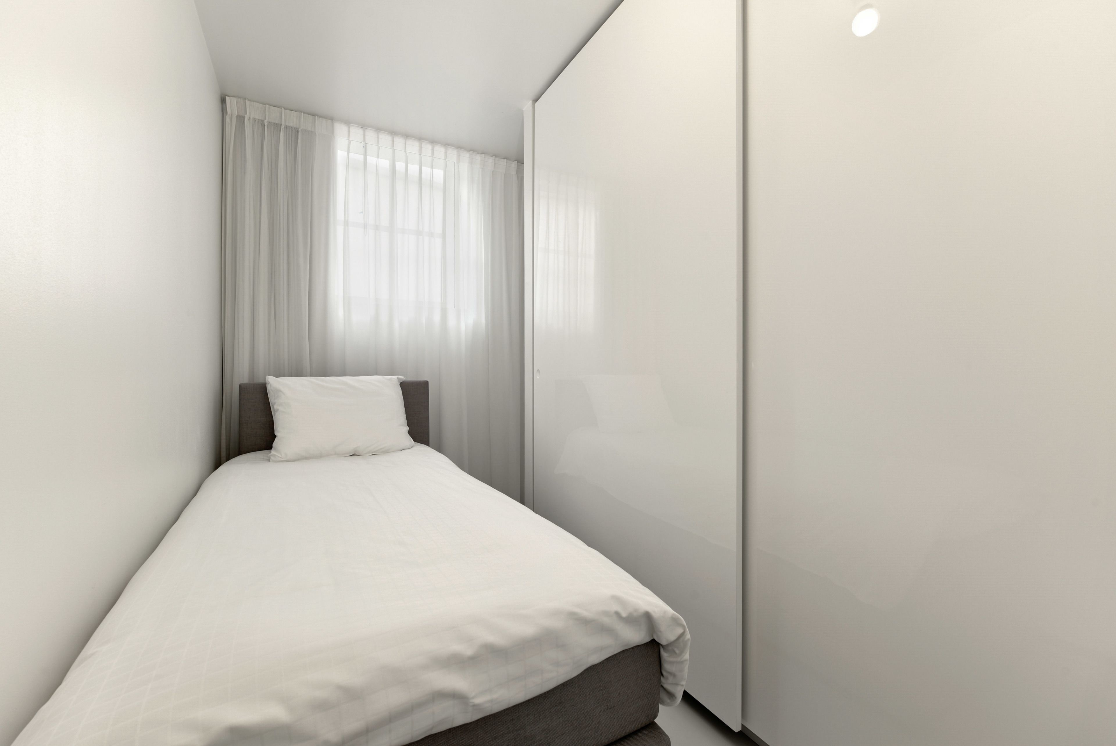 Ein kleines, ordentlich bezogenes Bett mit weißer Matratze und grauem Kopfteil in einer Raumecke, mit weißen Wänden und einem großen Kleiderschrank.