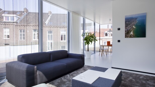 Una sala de estar moderna con un sofá oscuro, una mesa de centro blanca y un amplio ventanal con vistas a un paisaje urbano.