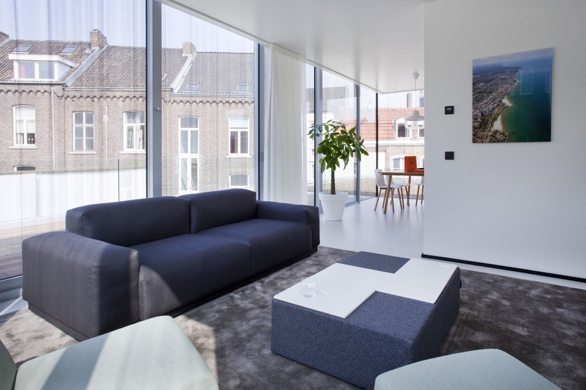 Ett modernt vardagsrum med en mörk soffa, ett vitt soffbord och ett stort fönster med utsikt över en stadsbild.