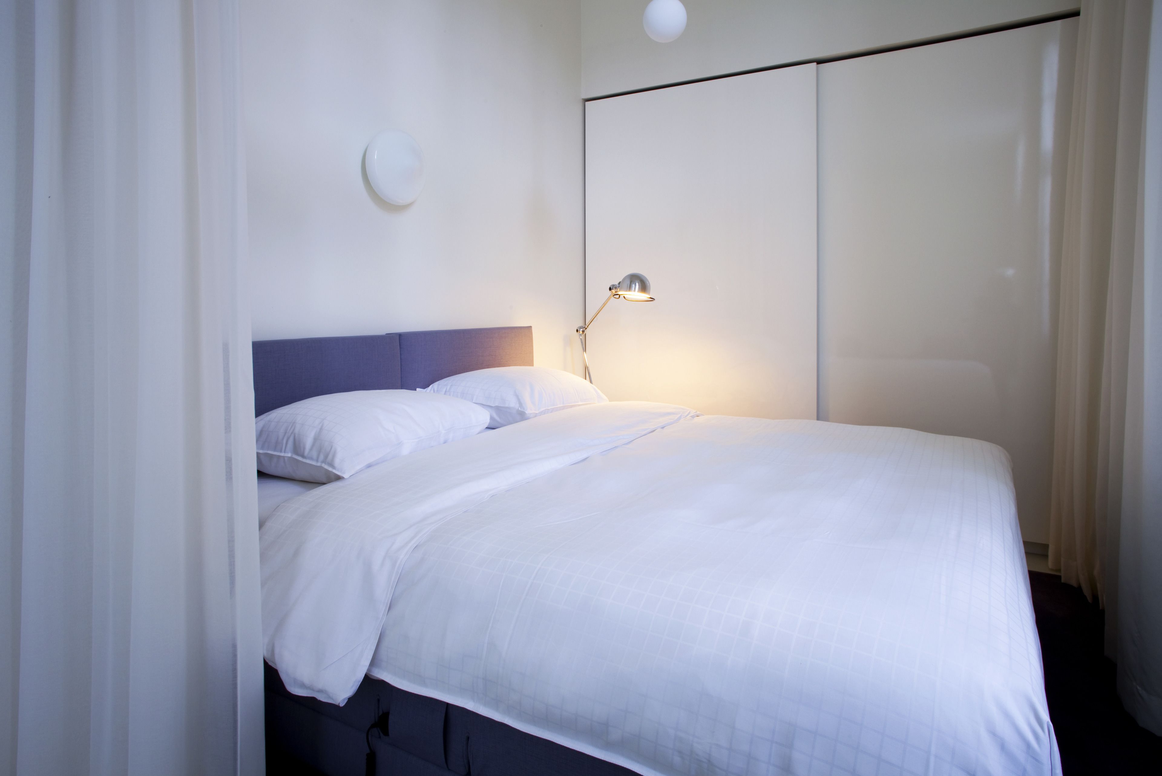Een net opgemaakt bed met witte lakens, een bedlampje en een wandlamp.