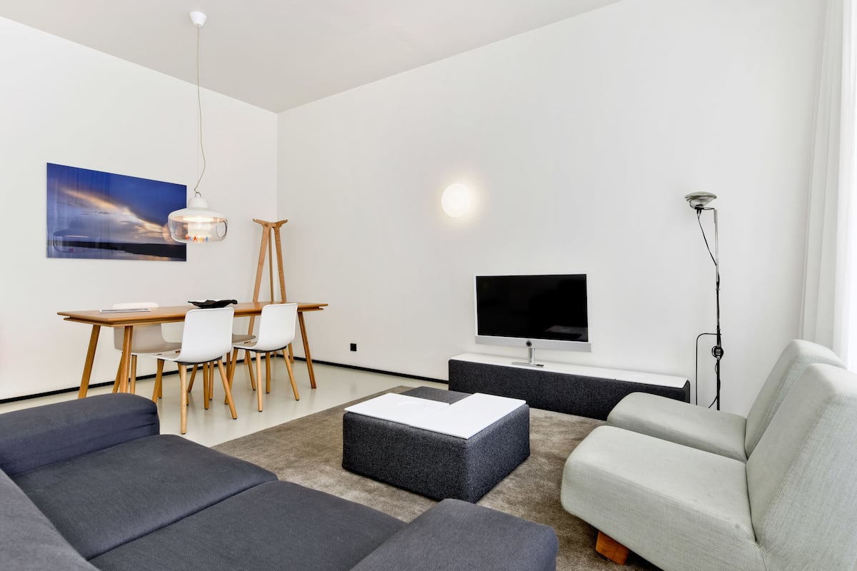 Ett modernt vardagsrum med en soffa, en pall, ett matbord, stolar, en tv och en tavla på väggen.
