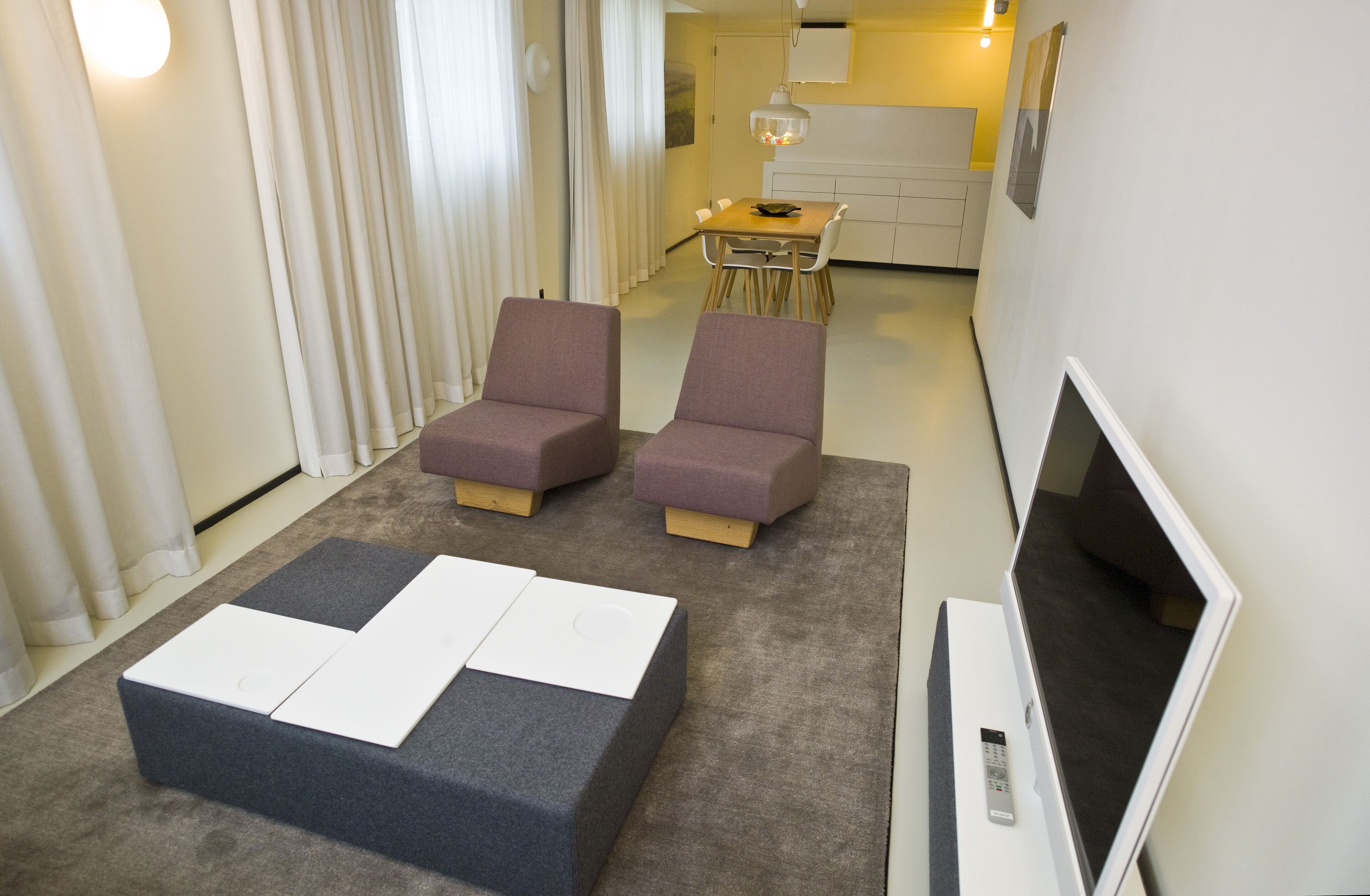 Una habitación de hotel moderna con sofá cama, televisor de pantalla plana y zona de comedor al fondo.