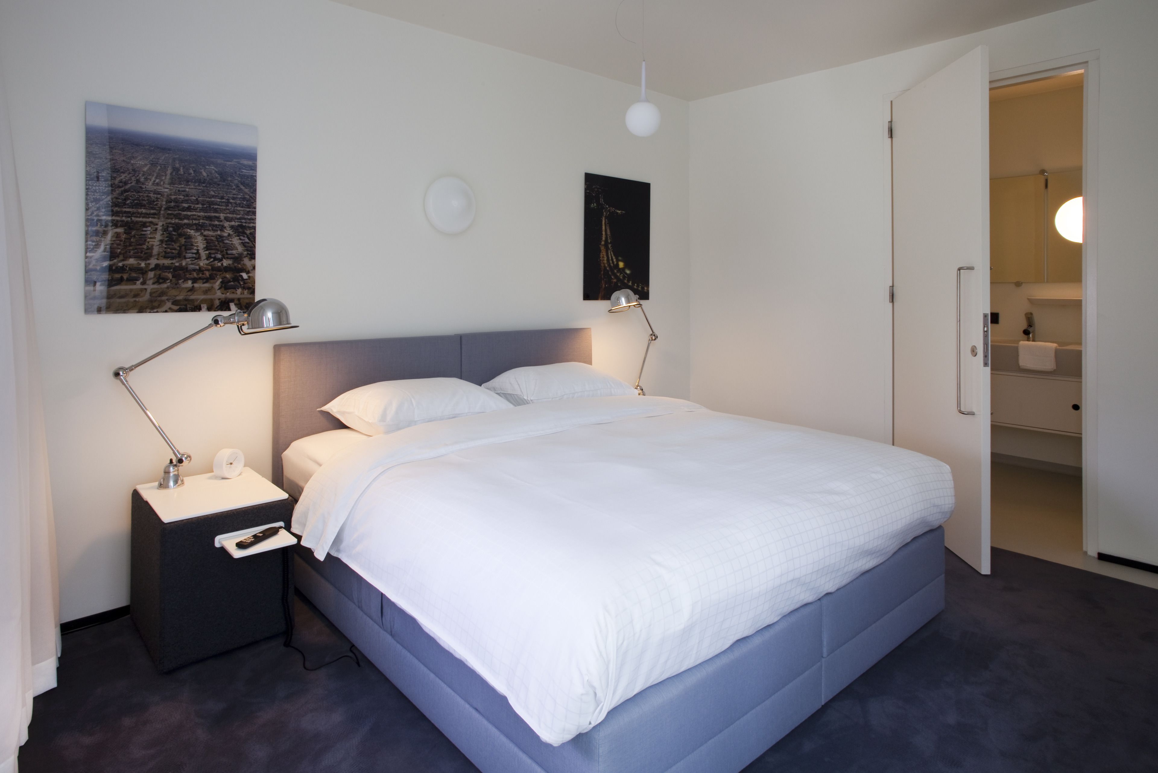Un dormitorio moderno con una cama grande, dos mesitas de noche con lámparas, una mesita auxiliar y una puerta que da a un baño.