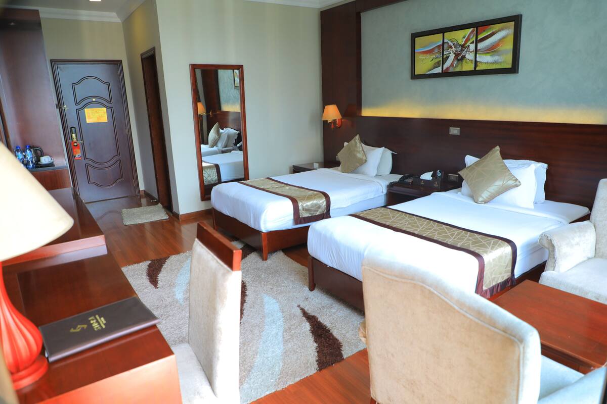 A hotel room with two beds, a desk, a chair, a lamp, and a painting on the wall.