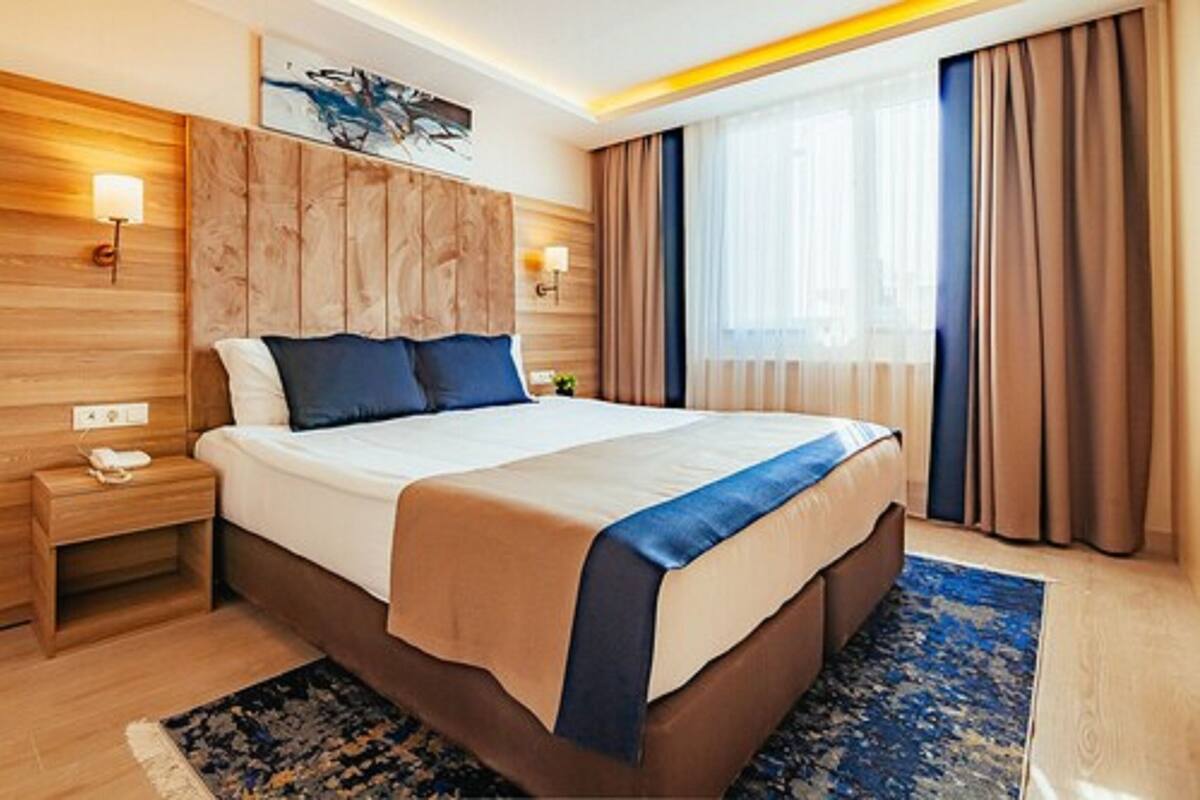 A hotel room with a large bed, wooden headboard, two bedside tables, a nightstand, and a window with curtains.