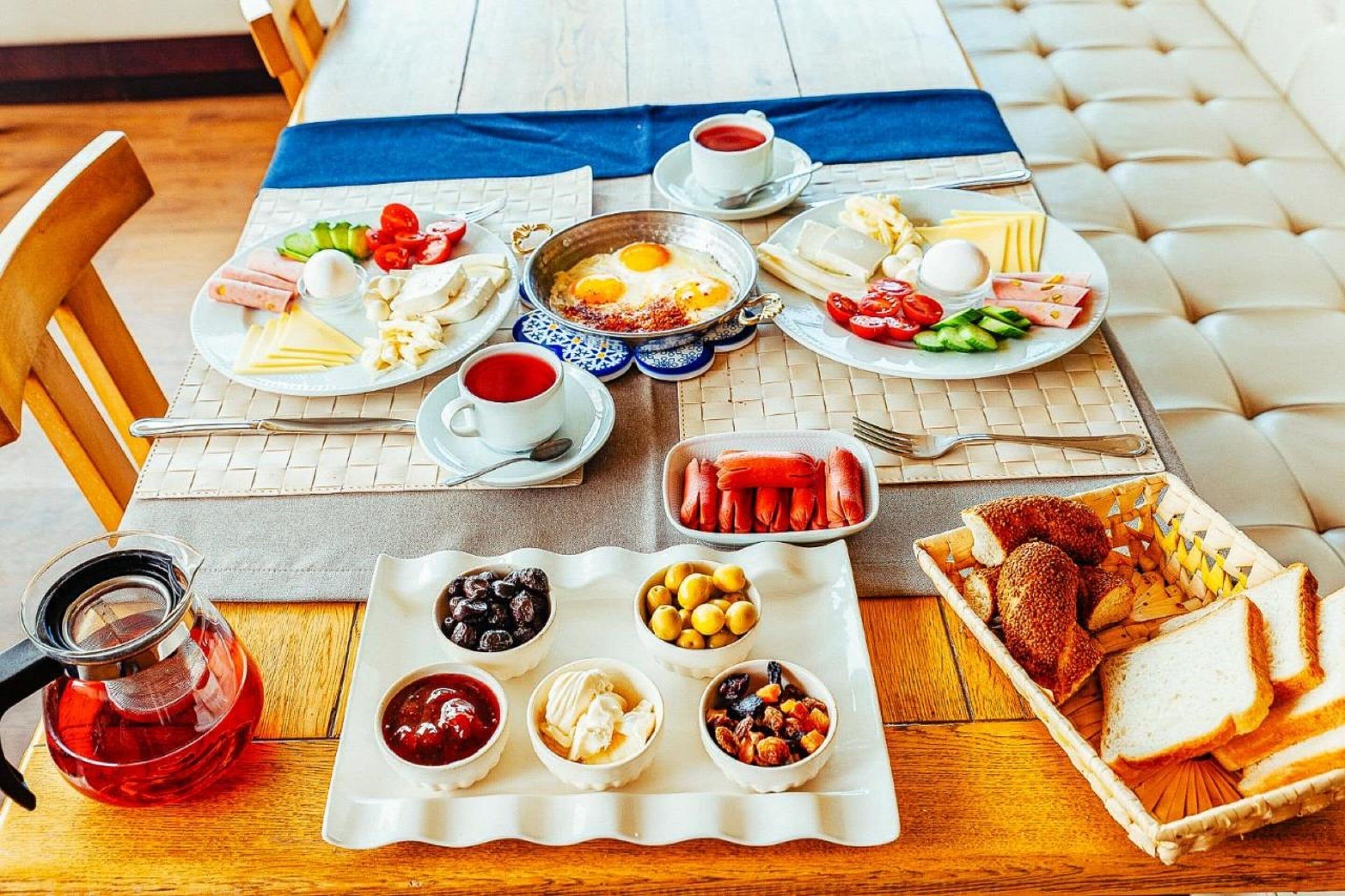 Une table mise pour le petit-déjeuner, avec des assiettes contenant des œufs, du fromage, des tomates et du pain, ainsi qu’une théière et des tasses.