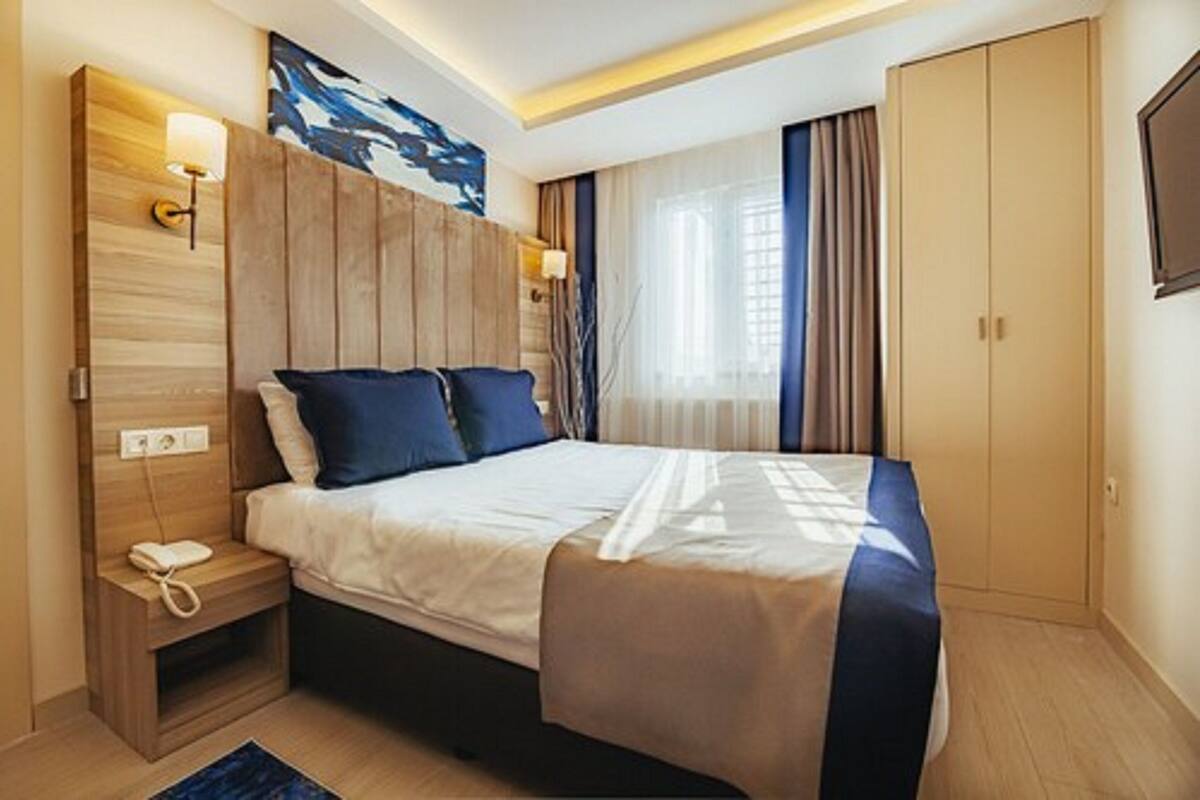 A modern hotel room with a large bed, wooden headboard, bedside lamps, a small bedside table with a phone, and a wardrobe.