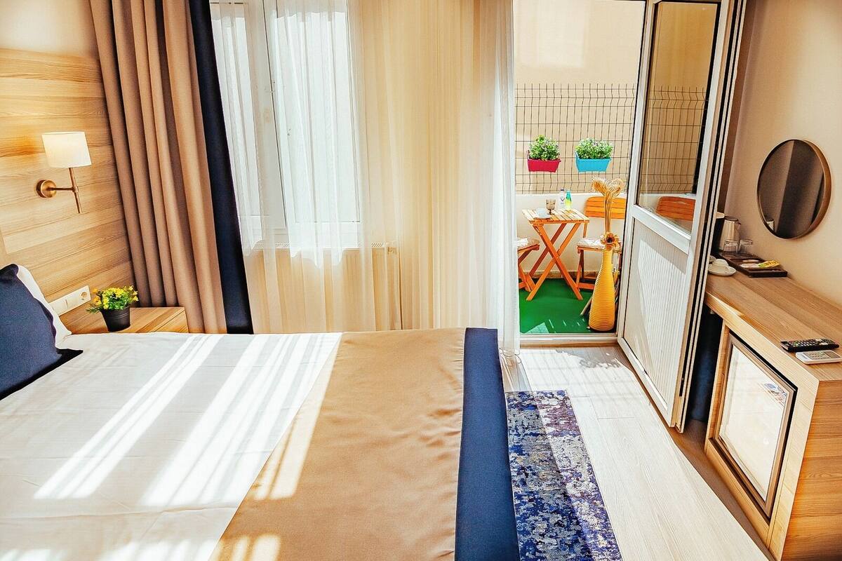 A hotel room with a bed, bedside table, lamp, curtains, and a view of the outdoor area with a table and chairs.
