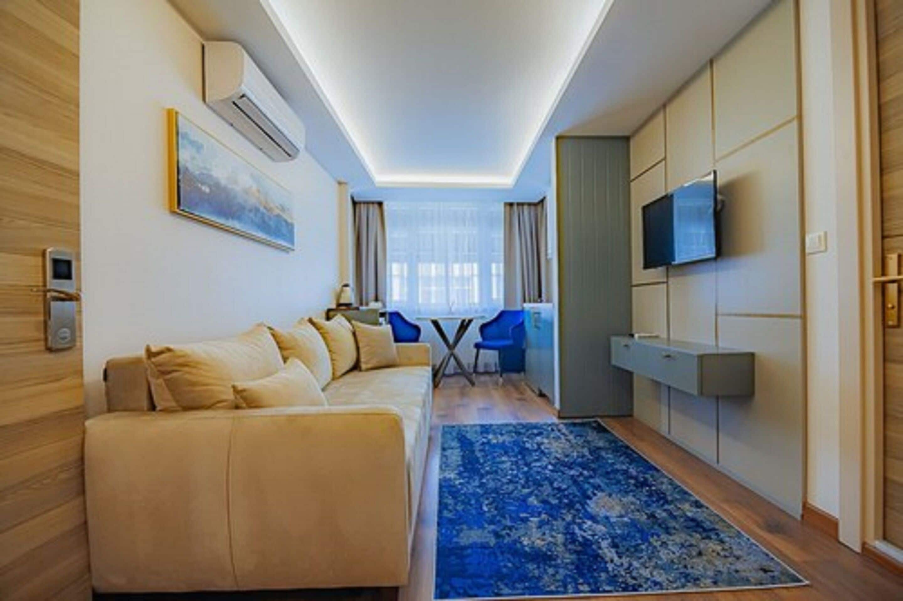 Una habitación de hotel moderna con sofá, alfombra azul, televisión montada en la pared y ventana con cortinas.