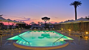 Una piscina con una isla central, rodeada de sillas de descanso y sombrillas, con el cielo de la puesta de sol de fondo.