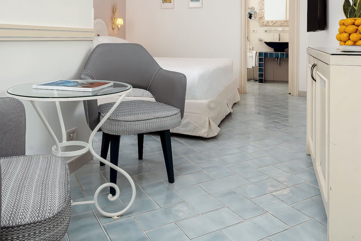 Una habitación de hotel moderna con una cama, una mesita con un libro, una silla, un sofá, y un baño visible a través de una puerta abierta, además de un televisor montado en la pared.