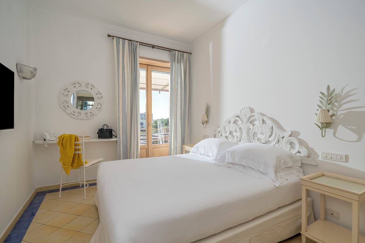 Un dormitorio con cama, mesita de noche, una silla, espejo, ventana con cortinas y vistas al exterior.