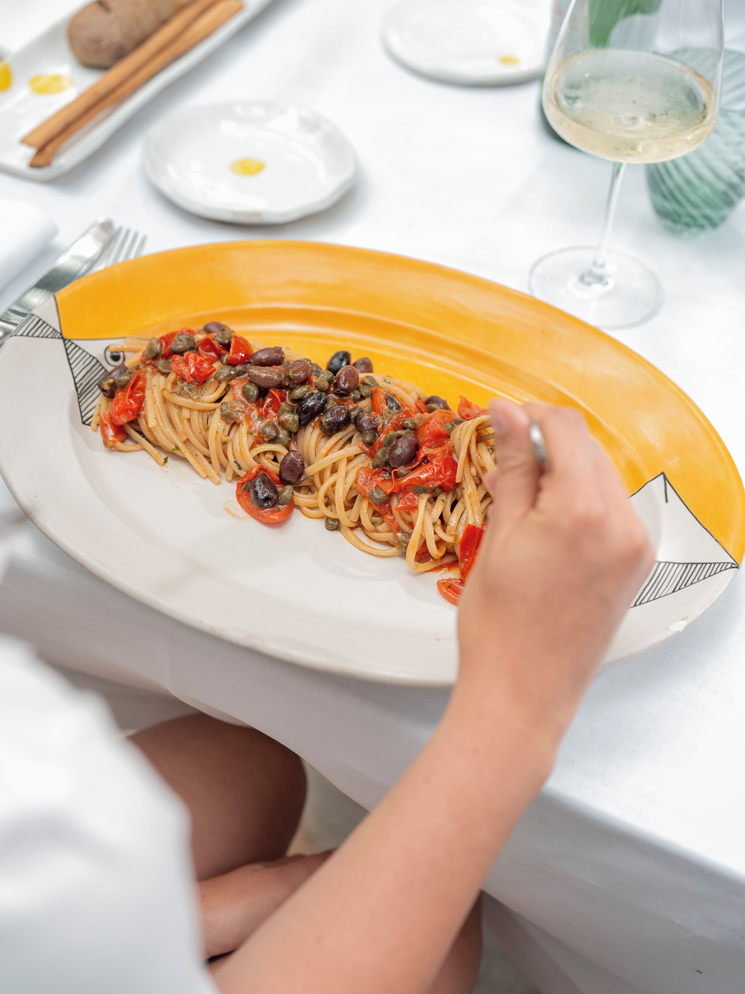 Un plato de espagueti con alcaparras y tomates, una copa de vino y un tenedor sobre una mesa.