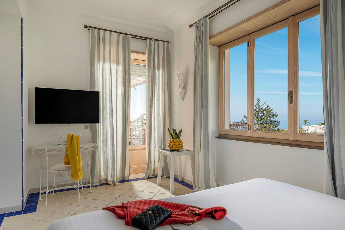 Habitación de hotel con cama, televisor, balcón con cortinas y vista al mar.