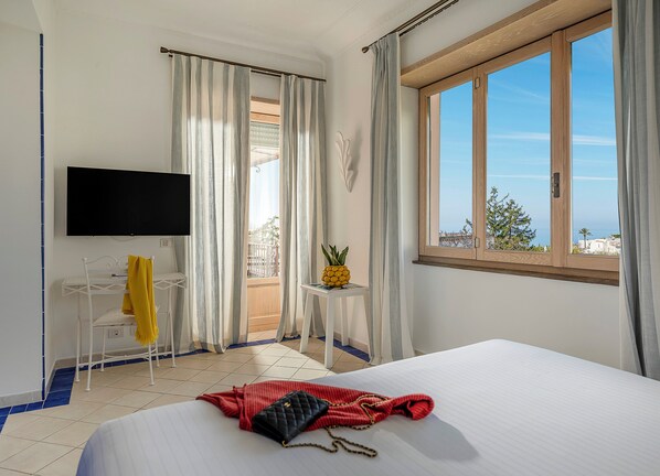 Habitación de hotel con cama, televisor, balcón con cortinas y vista al mar.