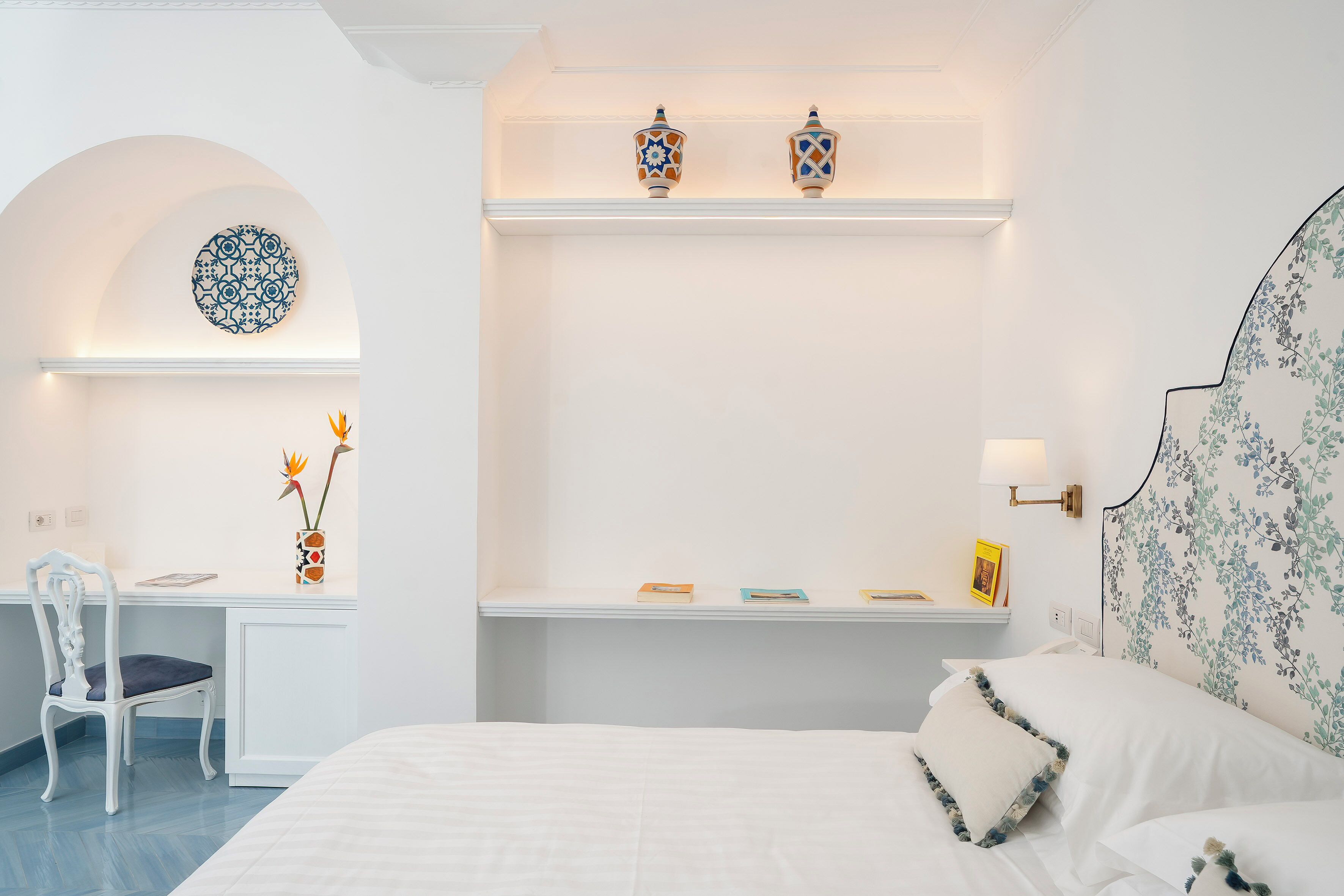 A bedroom with a bed, a desk with a chair, a shelf with decorative items, and a wall with a floral pattern.