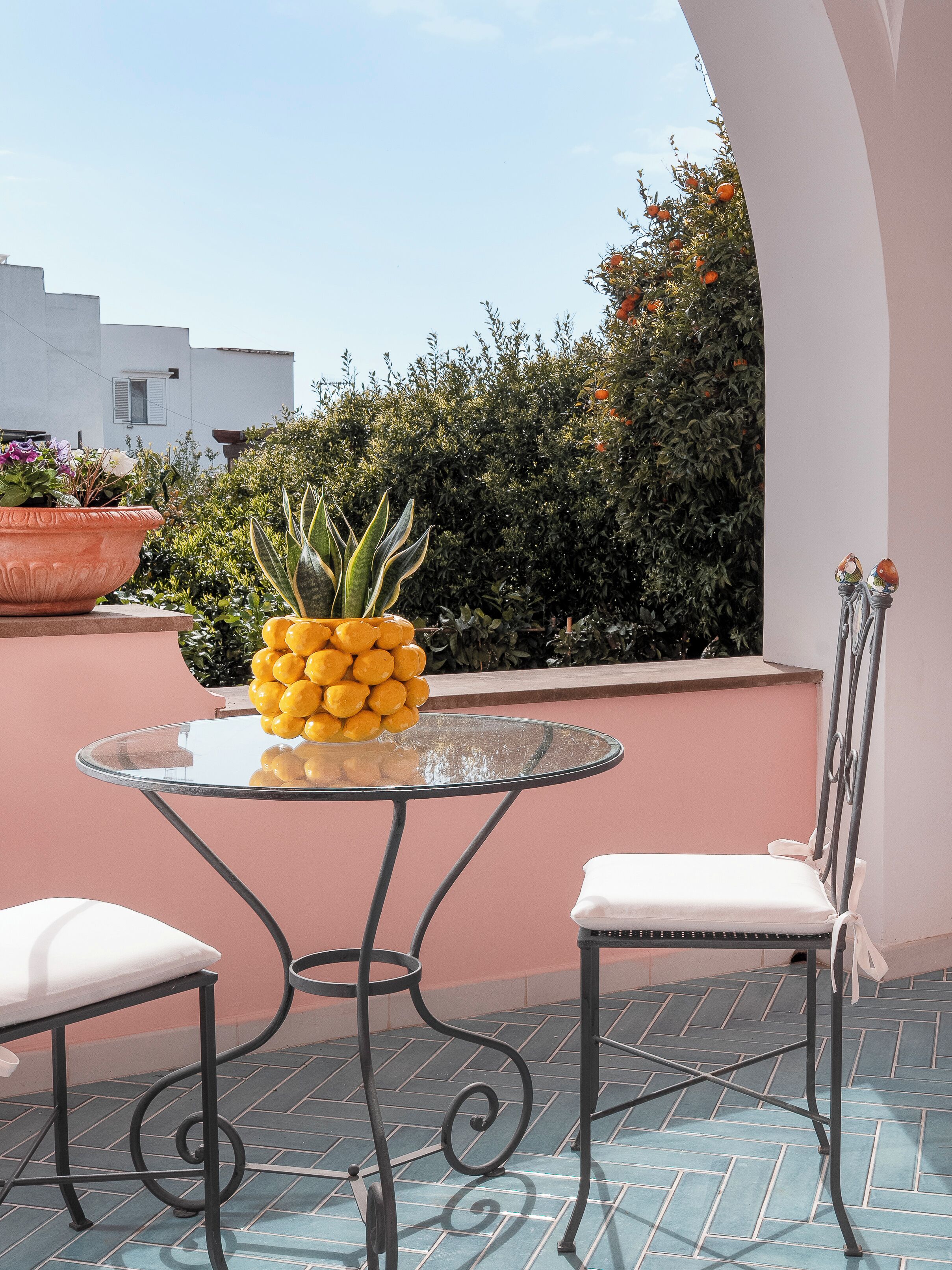 A glass table with a metal frame, two white chairs, a potted plant with yellow flowers, and a hedge in the background.