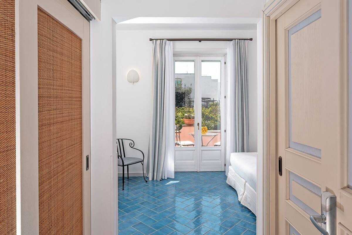 Una habitación con cama, una silla, un balcón con plantas y una puerta.