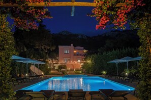 A swimming pool with lounge chairs and umbrellas, surrounded by greenery and a building in the background. - Casa Mariantonia (Anacapri)