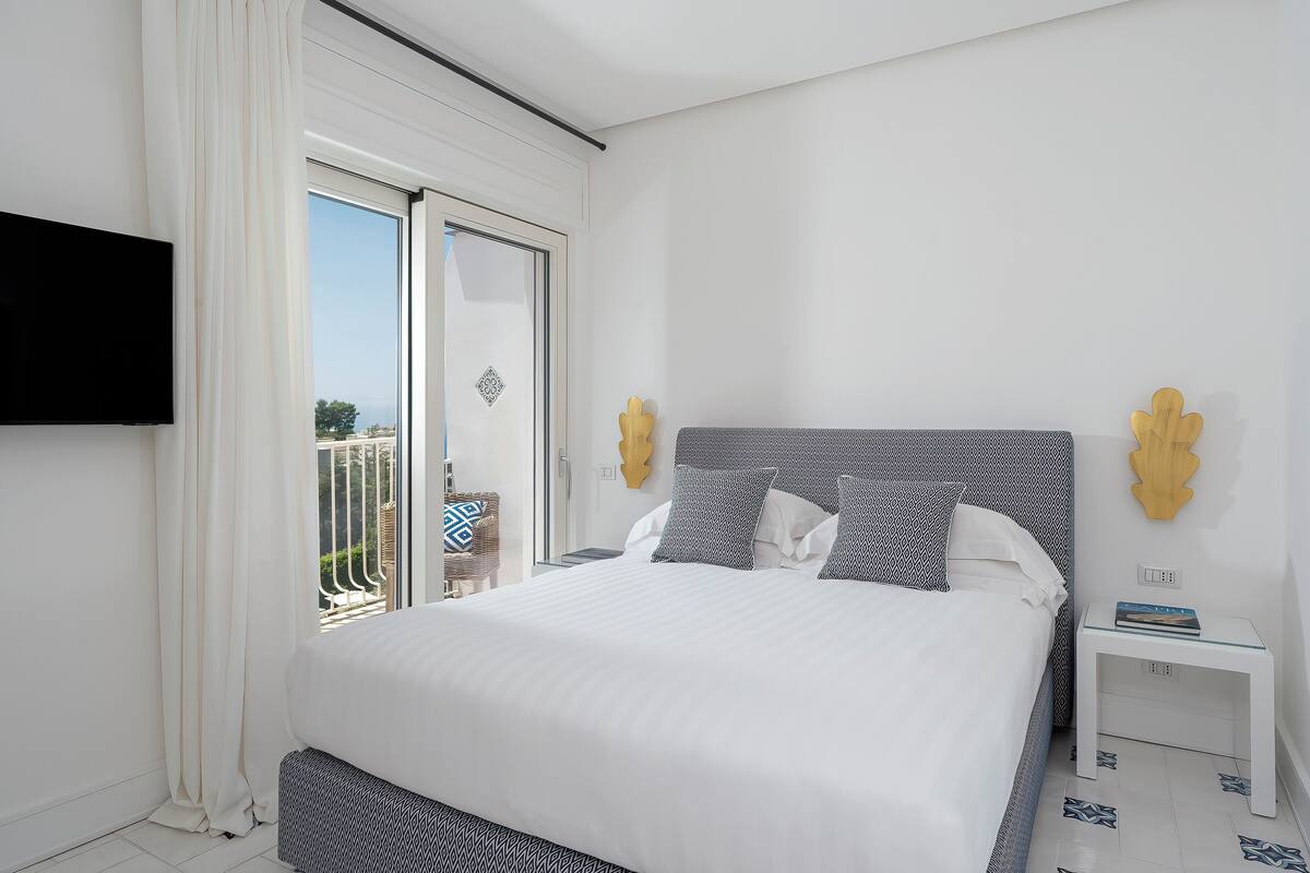 Un dormitorio moderno con una cama grande, una mesita de noche, un televisor montado en la pared y un ventanal corredero que da a un área exterior.