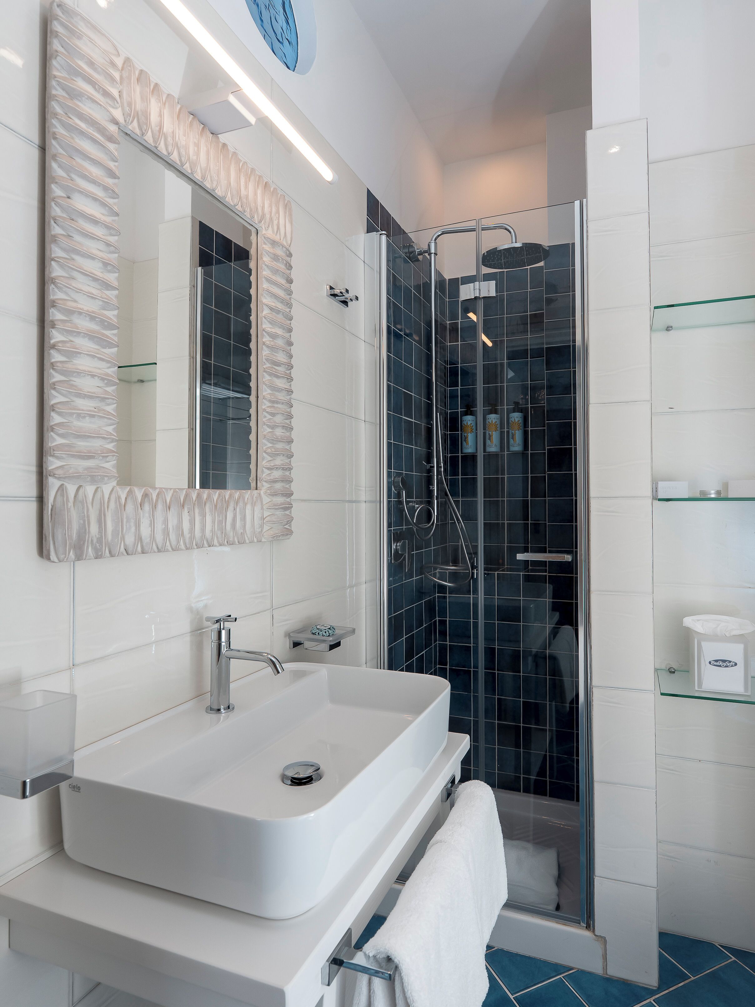A modern bathroom with a white sink, a large mirror, a glass shower enclosure, and a glass shelf with toiletries.