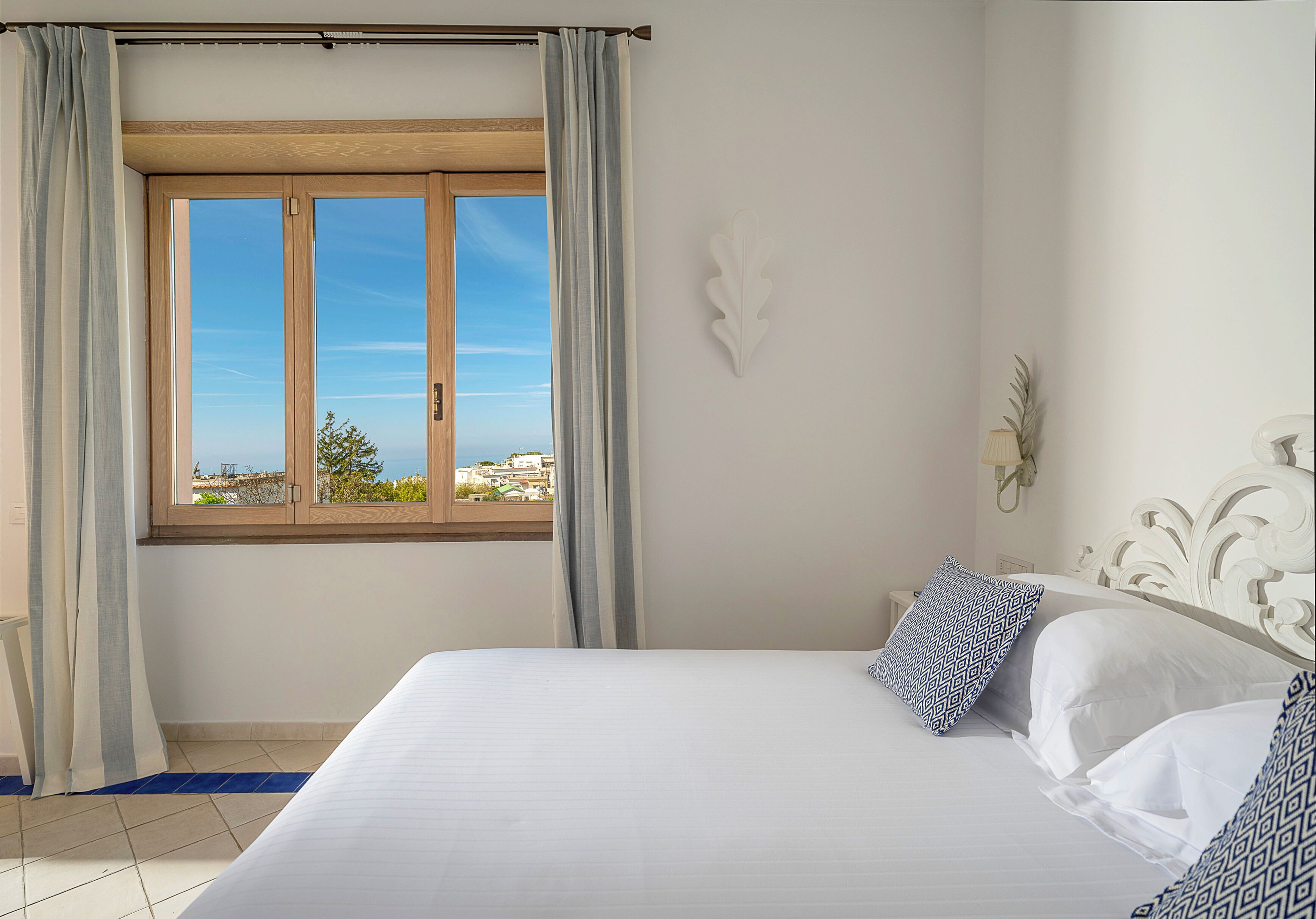 A bedroom with a large bed, a window with curtains, a decorative wall piece, and a view of buildings outside.