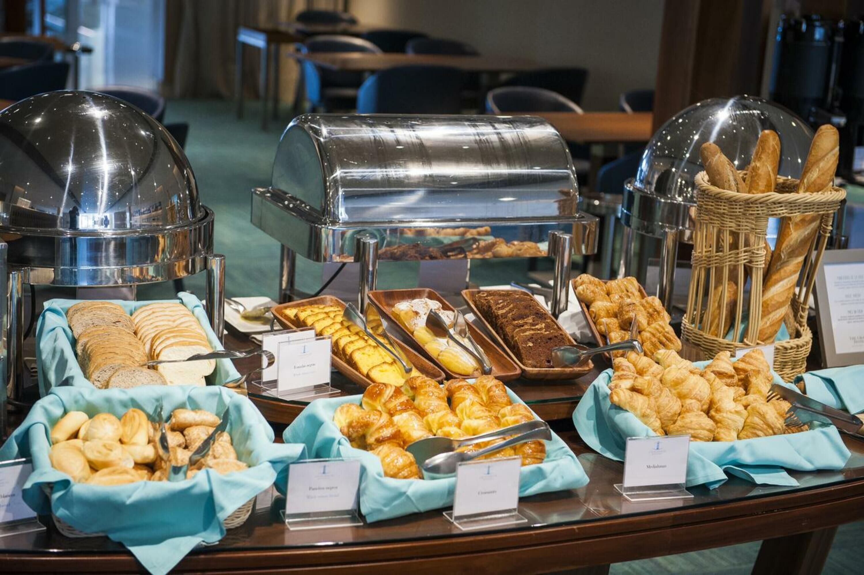 A buffet with various baked goods and pastries, including bread, croissants, and muffins, displayed in clear containers with labels.