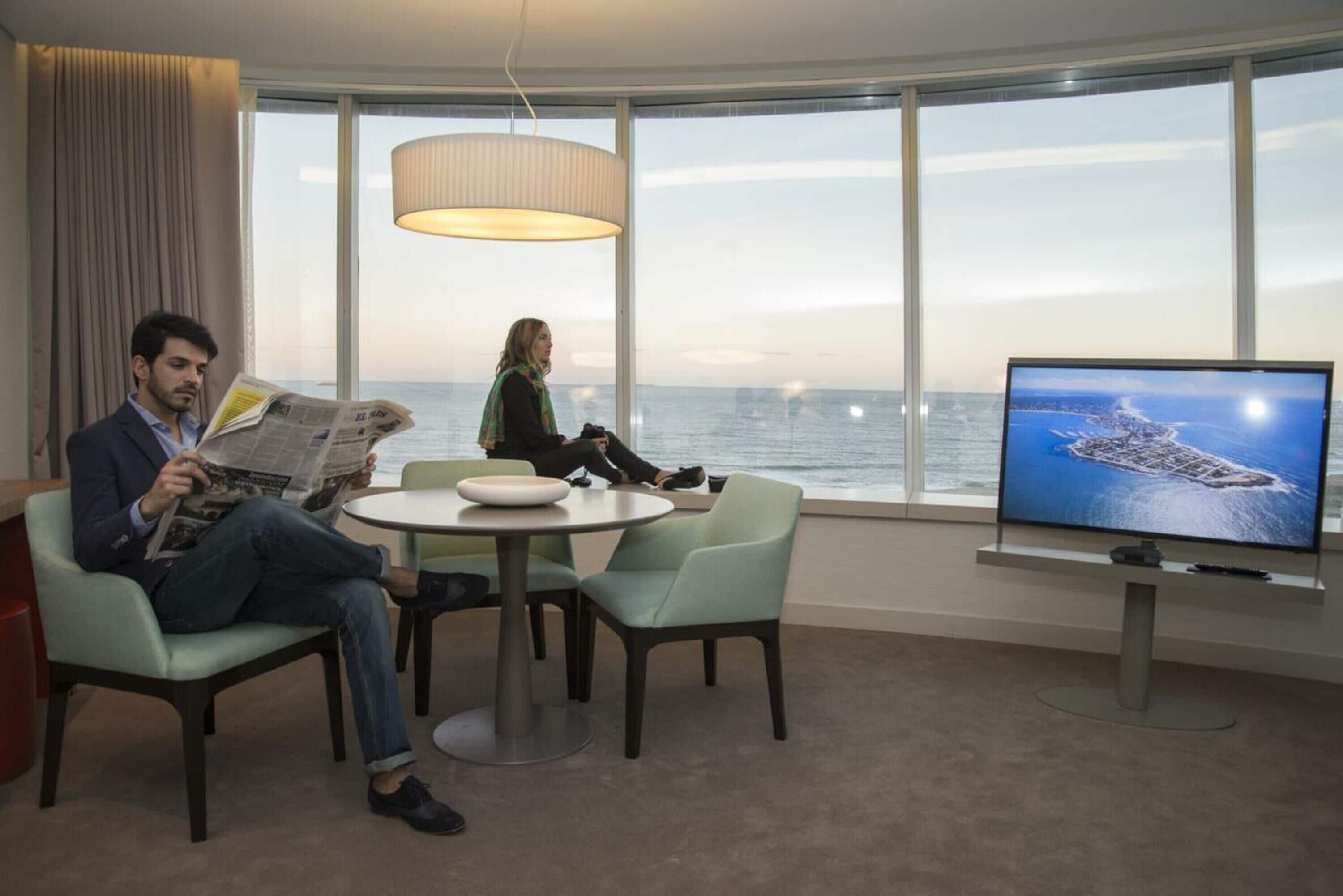 A room with a large window, a round table, two chairs, a television, and a person reading a newspaper.
