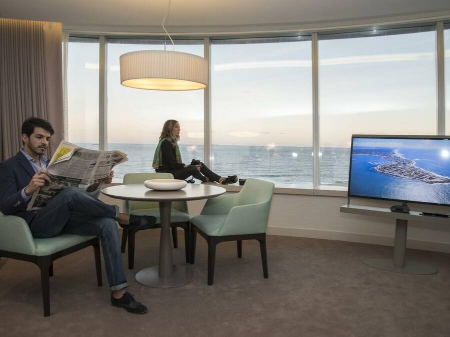 A room with a large window, a round table, two chairs, a television, and a person reading a newspaper.