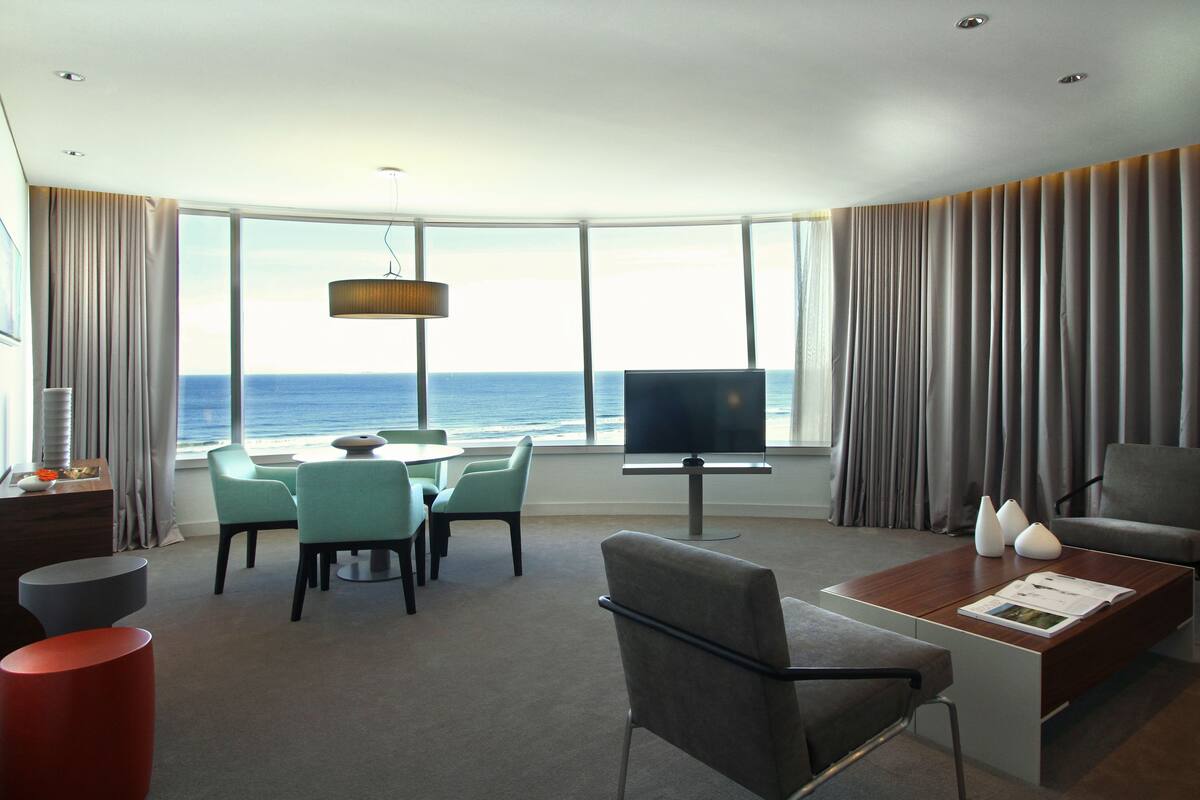 A modern hotel room with a large window offering a sea view, a flat-screen TV, a dining area with chairs, a coffee table, and a desk with a lamp.