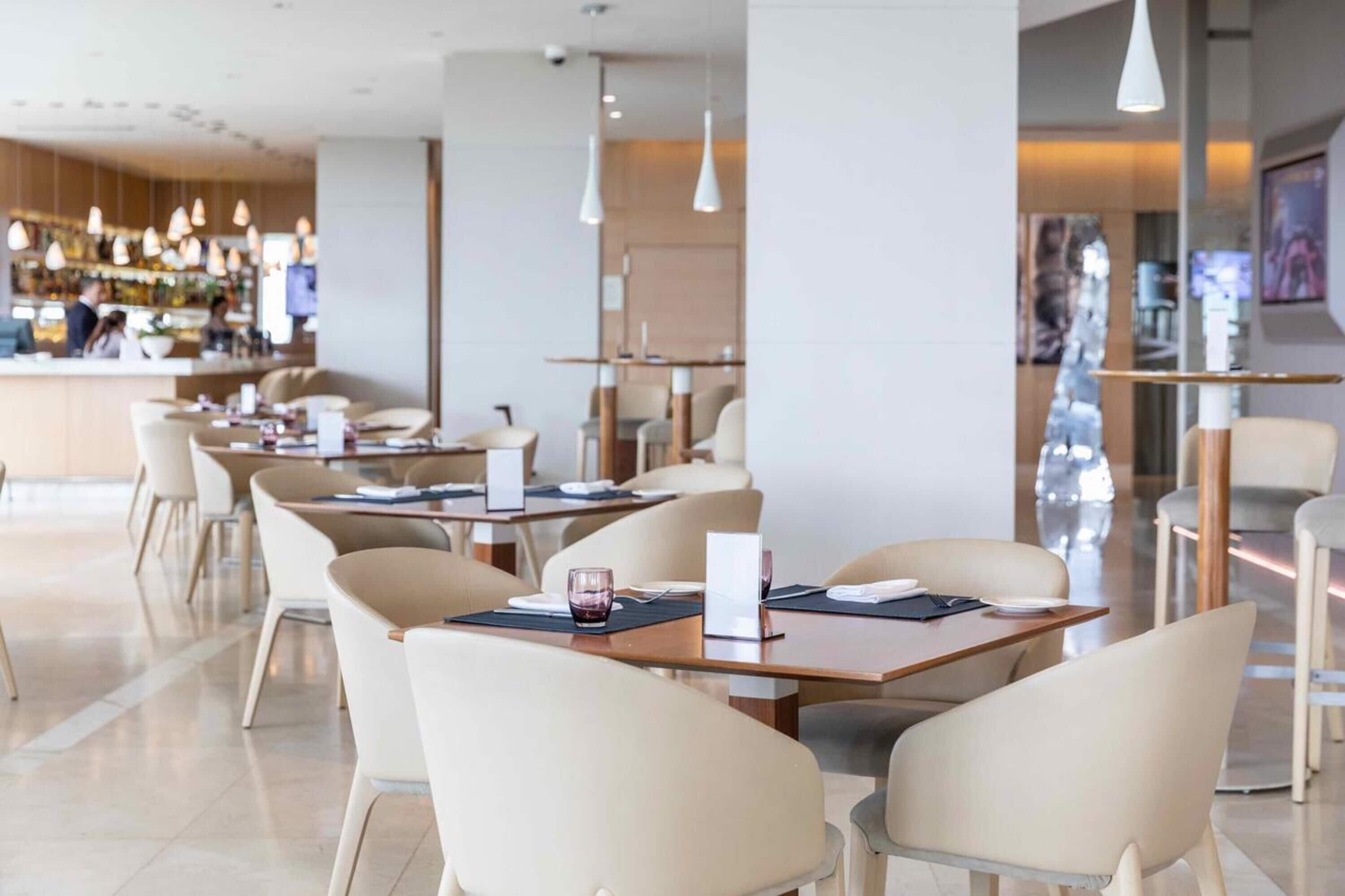 Un restaurante moderno con una zona de bar al fondo. Las mesas están dispuestas en línea, con sillas.
