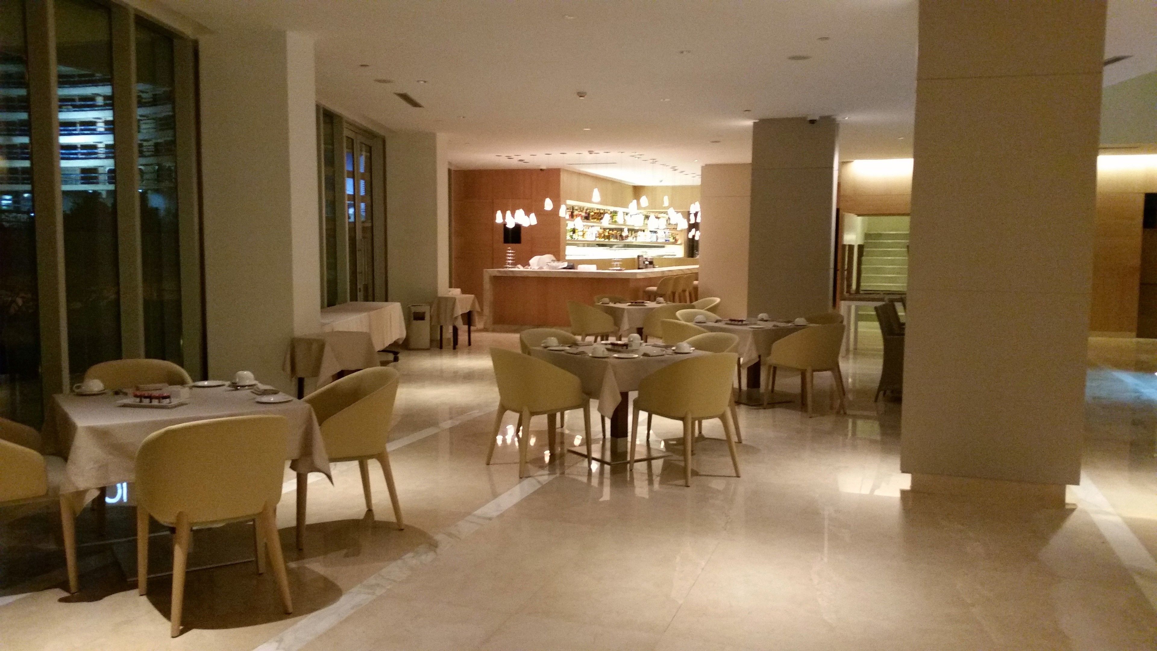 Uma área de refeições de hotel com várias mesas e cadeiras, um bar ao fundo e grandes janelas.