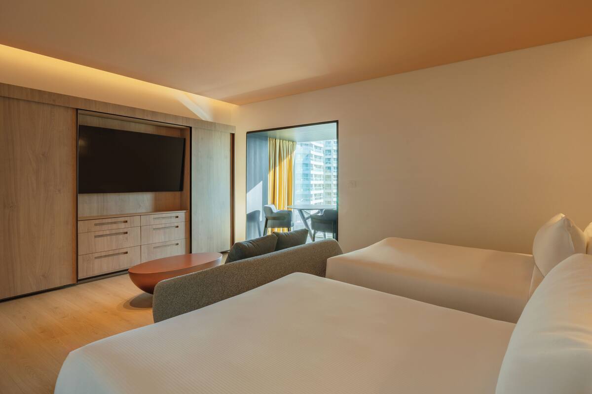 A modern hotel room with a large flat-screen TV, a comfortable sofa, two beds, and a view of the cityscape through a large window.
