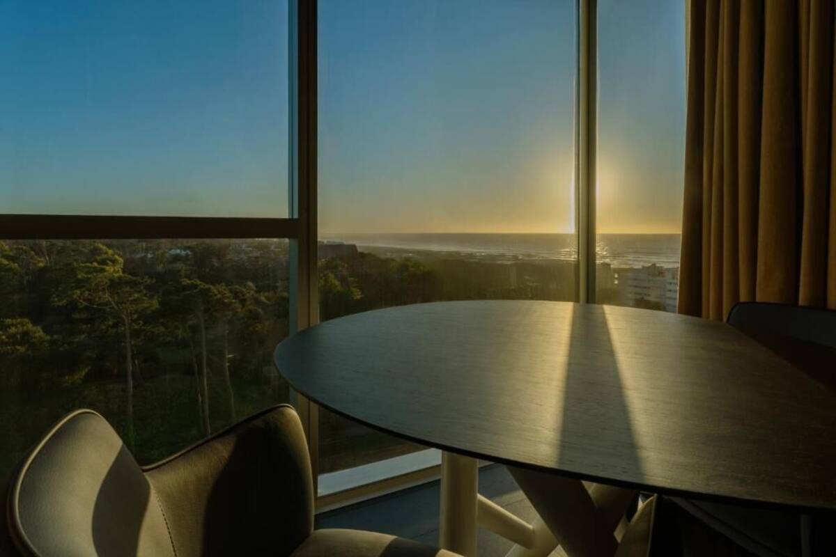 A room with a round table and chairs, large windows with a view of a cityscape and trees.