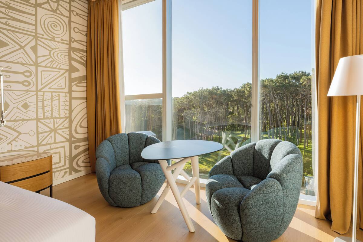 A modern hotel room with a large window, two armchairs, a small table, and a bed.
