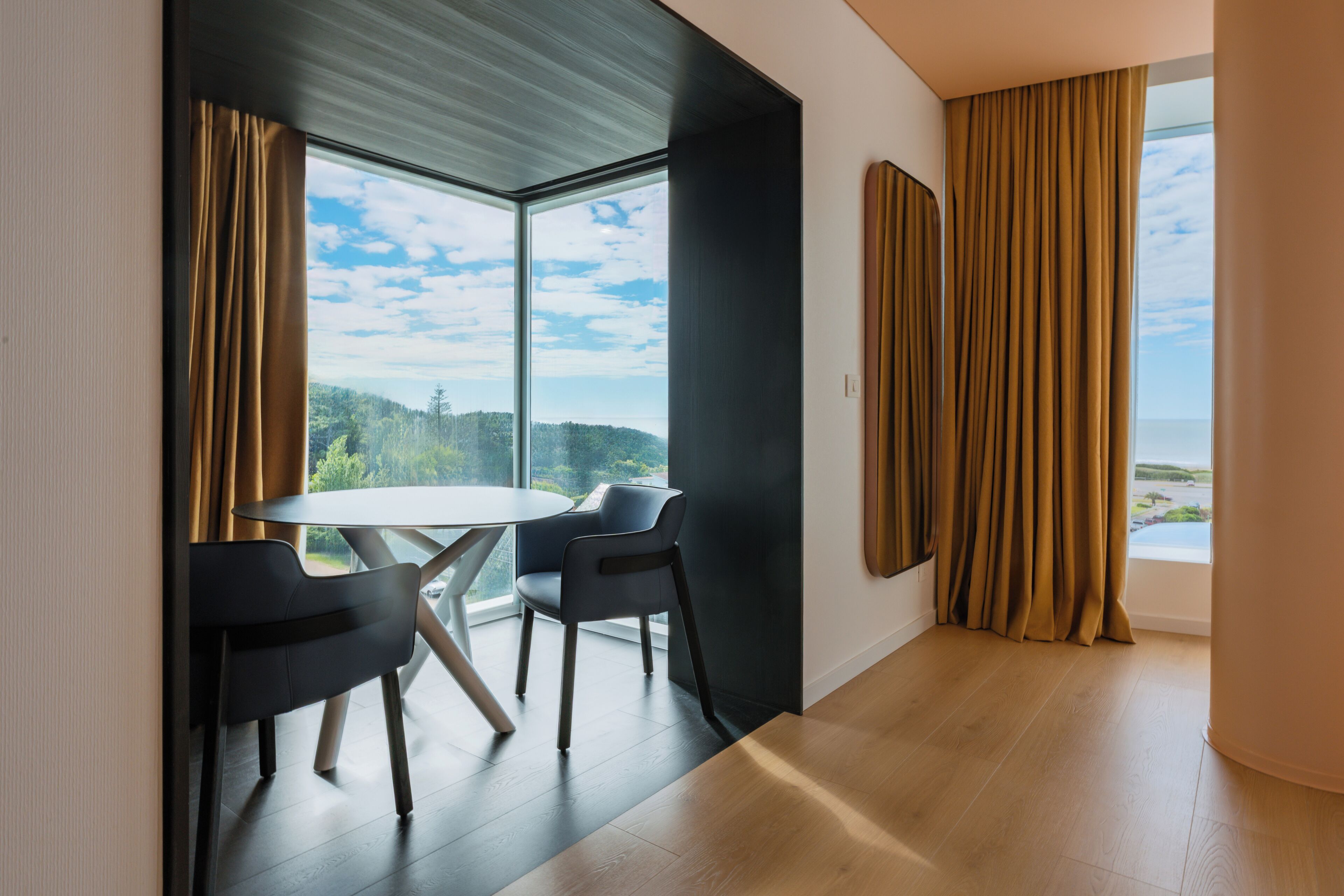 Una habitación moderna con una mesa redonda y dos sillas, ventanas amplias con cortinas y vistas a un área boscosa.